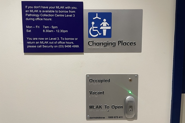 Close up of the closed door of the Changing Places facility. It includes a metal plate with 'Changing Places' and the the Changing Places symbol on it. A blue sign with information about how to borrow a MLAK. There is a second metal place which shows whether the facility is occupied or vacant and it has a lock on it.